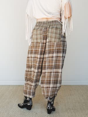 Shot from behind, this outfit pairs relaxed plaid barrel-leg pants in warm brown tones with a cream fringe top and embroidered black ankle boots — effortlessly lived-in with a boho-western edge.