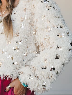 A close-up detail shot of a cream feathered jacket scattered with oversized gold and silver sequins — the kind of piece that makes a regular Tuesday feel like a celebration.