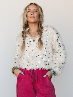A fuzzy cream sweater scattered with gold and silver sequins is paired with deep magenta wide-leg joggers — the kind of outfit that feels festive without trying too hard, styled with layered gold necklaces and stacked boho bracelets.