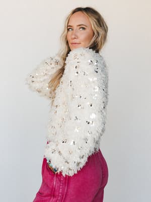 A model looks over her shoulder in a cream fluffy sequin sweater paired with deep magenta jeans — the kind of outfit that makes a Tuesday feel like a reason to get dressed.