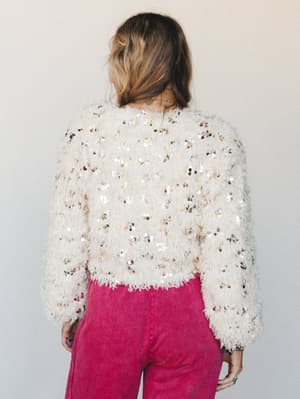 Shot from behind, a model wears a cream fluffy cropped jacket covered in scattered silver and gold sequins, paired with faded hot pink wide-leg pants — the kind of outfit that walks into a room and doesn't need to say a word.