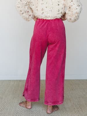 A back-view shot of wide-leg acid-wash pants in a rich berry-pink paired with a fluffy sequin-embellished cream sweater, styled barefoot on a woven rug for an effortlessly expressive, lived-in feel.