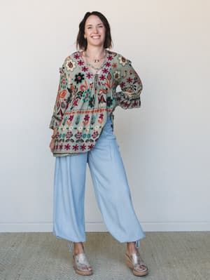 A smiling model wears a flowy sage floral print top with wide-leg light wash pants and metallic wedge sandals — the kind of put-together-but-relaxed outfit that looks like it came together effortlessly.