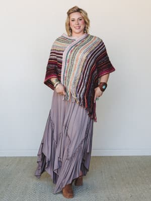A smiling blonde model layers a chunky multicolor striped knit poncho over a flowy dusty mauve maxi skirt with tassel trim details, styled with stacked boho bracelets and tan block-heel boots — the whole look reads like a fall farmers market in the b