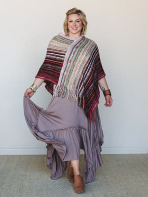 A smiling model in a flowy mauve maxi skirt and a richly striped multicolor knit poncho with fringe — the kind of layered, expressive outfit that feels like it was assembled with intention and a good eye.