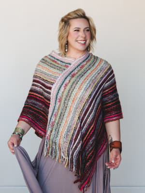 A smiling blonde model wears a richly striped multicolor knit poncho with fringe trim over a flowing mauve wide-leg pant, styled with layered bracelets and drop earrings — warm, lived-in, and full of personality.