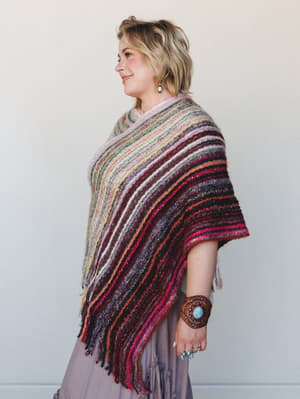 A side-profile shot of a model wearing a richly striped, fringed knit poncho layered over a dusty mauve midi skirt, styled with a leather turquoise cuff and stacked rings — the kind of outfit that looks like it traveled somewhere interesting.