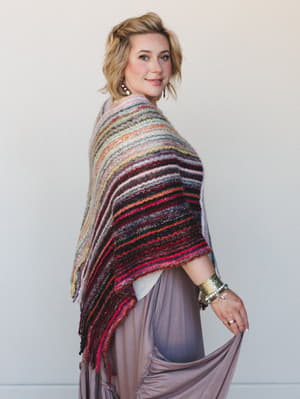 A warm, lived-in shot of a model draped in a richly striped multicolor knit wrap — all fringe edges and layered texture — paired with a flowy mauve maxi skirt and stacked boho jewelry that feels like a whole mood in the best way.