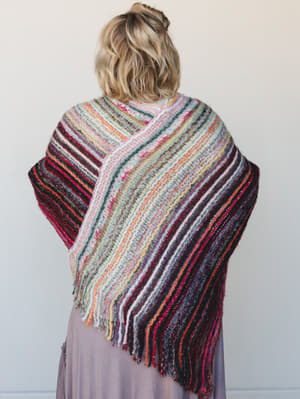 Shot from behind, a model drapes a richly striped multicolor knit wrap over her shoulders, its diagonal fringe hem falling against a soft mauve skirt — the kind of piece that looks like it has a story.