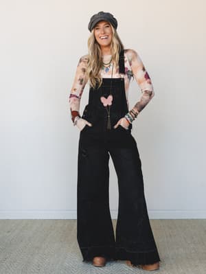 A smiling blonde model in wide-leg black denim overalls layered over a long-sleeve floral mesh top, styled with a charcoal newsboy cap, brown clogs, and stacked boho jewelry — the kind of outfit that looks like it came together effortlessly but defin