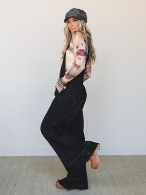 A side-profile shot of a model in wide-leg black denim overalls layered over a long-sleeve floral top, styled with a charcoal baker boy cap and stacked boho bracelets — the kind of outfit that looks effortless but was absolutely thought about.