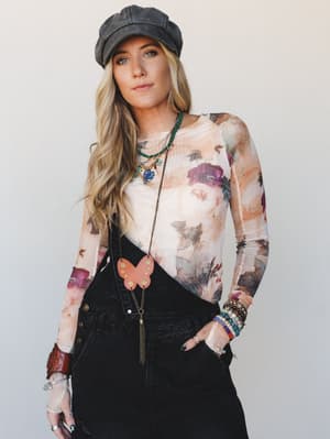 A layered boho look built around a sheer floral mesh long-sleeve top tucked into black denim overalls, styled with a faded newsboy cap, stacked beaded bracelets, and a long butterfly pendant necklace — the kind of outfit that looks effortless but cle