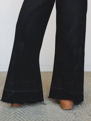 A close-up of wide-leg black denim jeans with a dramatic flare and raw frayed hem, styled with tan wooden clogs on a woven jute rug — the kind of detail shot that makes you understand exactly how good these are going to look in person.