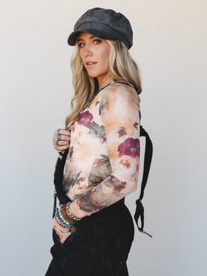 A blonde model shot in three-quarter profile against a soft grey studio wall, wearing a sheer floral long-sleeve top layered over what appears to be a bodysuit, paired with black cargo pants and a washed charcoal newsboy cap — the whole look has that