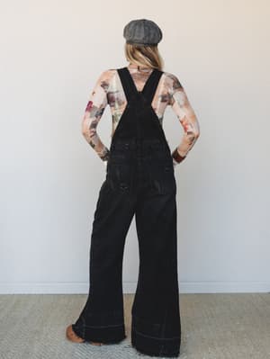 Shot from the back, this look pairs wide-leg black denim overalls with a floral mesh long-sleeve top — a lived-in, expressive combo that feels effortlessly vintage and a little artsy, finished with a newsboy cap and stacked bracelets.