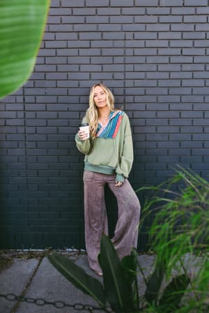 A relaxed, coffee-in-hand moment — she's leaning against a dark brick wall in an oversized sage sweatshirt with colorful patchwork detailing and wide-leg mauve pants, framed by tropical greenery like the city paused just for her.