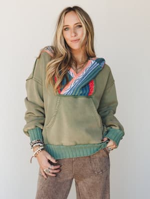 An olive green washed hoodie with a bold serape-stripe hood lining gets styled with acid-wash wide-leg pants and a full arm of stacked bracelets — cozy and expressive in equal measure.