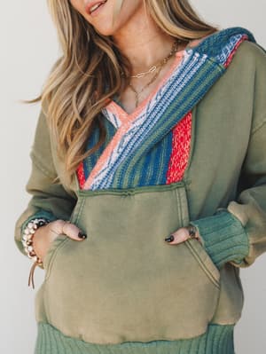 A close-up of a washed olive hoodie with a bold serape-stripe knit panel across the chest and hood, styled with layered gold necklaces, stacked rings, and a beaded bracelet — the kind of piece that feels like a weekend personality.