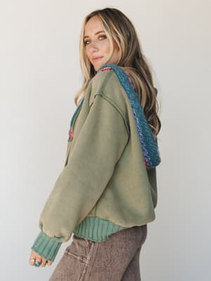 A model looks back over her shoulder wearing a washed sage green sweatshirt with a colorful chunky knit hood, paired with dusty mauve wide-leg jeans — the kind of effortless layered look that feels equally at home on a coffee run or a slow weekend mo
