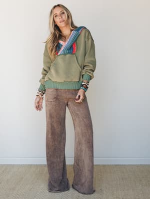 An olive green oversized hoodie with colorful woven patchwork detailing at the hood is paired with wide-leg mineral-washed mauve pants, styled with stacks of beaded bracelets and rings for a relaxed, expressive boho feel.