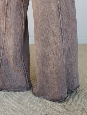 A close-up detail shot of wide-leg pants in a moody mauve-brown mineral wash, showing the relaxed drape and raw hem edge resting on a woven jute rug.