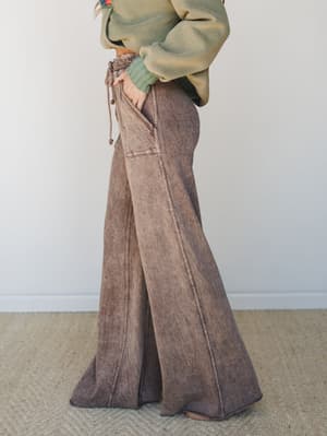 A side-profile shot focused on dramatically wide-leg, mineral-washed flare pants in a warm mocha-brown paired with a sage green oversized sweatshirt — the kind of effortless combo that looks like you tried just enough.