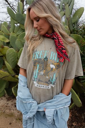 A woman with long blonde hair stands in front of cacti, wearing the Fly Free Graphic Tee - Mocha, a red patterned scarf, and a light denim jacket draped over her arms, looking down thoughtfully.