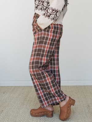 A side-profile shot of wide-leg plaid pants in warm brown, rust, and cream, styled with a fair isle knit sweater and tan studded clogs — the kind of autumn outfit that feels like it came together on purpose but also just happened.