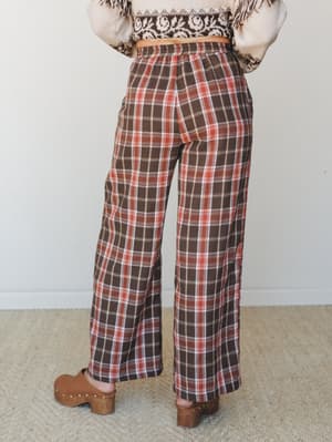 A back-view shot of wide-leg plaid pants in warm chocolate and rust, styled with a cream fair isle sweater and wooden clogs — the kind of autumn outfit that feels cozy without trying too hard.