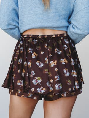 A flowy floral mini skirt in deep plum-brown is styled with a soft blue knit sweater — the kind of pairing that feels effortless and a little romantic, like she got dressed in five minutes and still looks like she tried.