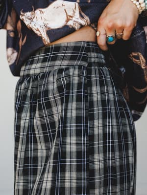 A close-up shot showing the waistband and hip of black plaid joggers styled with a cropped navy graphic tee — stacked silver rings and a turquoise statement ring on the model's hand pull the top up, giving the whole thing an effortlessly personal, co