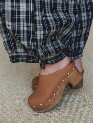 A close-up detail shot of plaid joggers with a gathered ankle tie, styled with tan studded clogs on a jute rug — earthy, lived-in, and the kind of pairing that feels effortless without trying.
