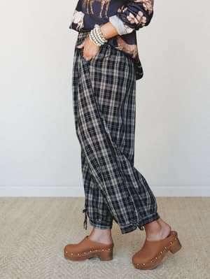 A side-angle shot of relaxed plaid joggers in dark charcoal and tan, styled with a floral graphic sweatshirt, stacked bead bracelets, and warm brown clogs — the kind of outfit that feels pulled-together without trying.
