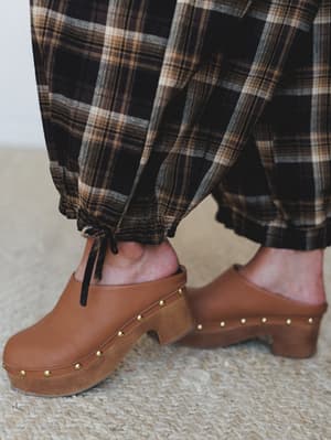 A close-up detail shot of wide-leg plaid pants with gathered, tie ankles paired with cognac leather platform clogs with gold stud trim — the kind of fall outfit that feels effortless and a little vintage all at once.