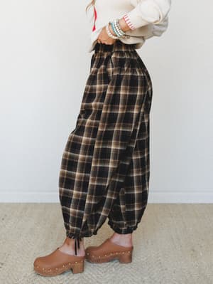 A side-profile shot of voluminous plaid harem-style pants in a dark espresso plaid, styled with a cream knit sweater, stacked bead bracelets, and studded tan clogs — the kind of outfit that feels like a cozy Saturday with a little personality.