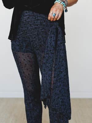 A close-up of navy crushed velvet pants with a sheer polka dot overlay and a side tie detail, styled with a black velvet top and layered turquoise and stone jewelry — moody and a little unexpected.