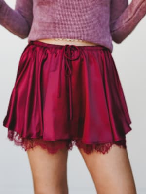 A satiny burgundy skort with a flirty flared silhouette, drawstring waist, and lace-trimmed hem layered beneath — paired with a cropped mauve thermal top for a soft, romantic mood.