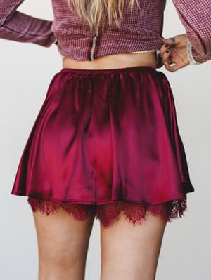 A close-up back view of a satin burgundy skort with a lace hem peeking out beneath, paired with a mauve waffle-knit crop top — the kind of outfit that feels equally at home at a holiday dinner or a late-night concert.