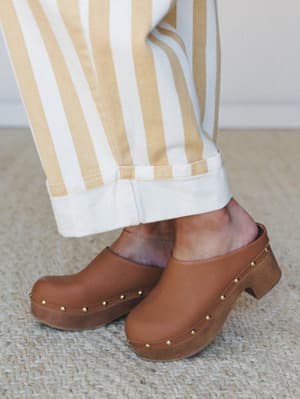 A close-up of camel leather clogs with gold stud trim and a wood-look platform heel, styled with wide-leg striped trousers in cream and warm gold — the kind of shoe-and-pant combo that makes the whole outfit feel intentional.