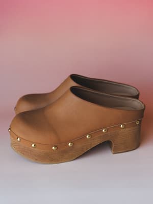 A pair of camel leather clogs with gold stud trim and a wood-textured platform heel, shot against a soft pink gradient — the kind of shoe that makes a whole outfit feel intentional.