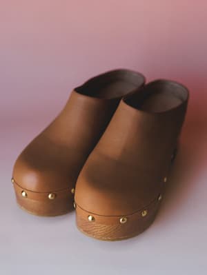 A pair of camel leather platform clogs with gold stud detailing along the wooden base — shot close against a soft pink-to-lavender gradient backdrop with a warm, editorial feel.