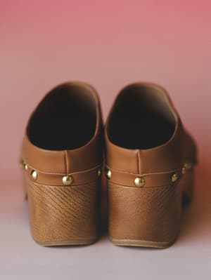 A close rear shot of camel leather clogs with gold stud trim along the heel strap, sitting against a soft pink backdrop — the kind of shoe that makes a whole outfit feel more intentional.