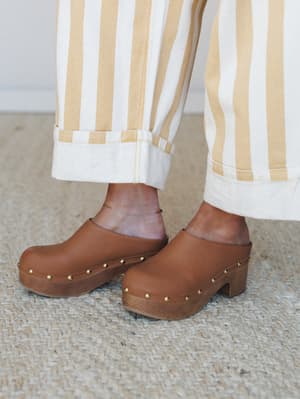 A close-up detail shot of tan leather clogs with gold stud trim and a wooden platform sole, paired with wide-leg striped trousers with a cuffed hem — earthy, effortless, and very much a 'yes to both' situation.