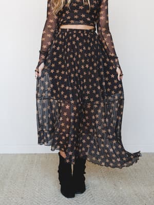 A sheer black maxi skirt covered in warm golden-brown star print floats mid-twirl, paired with a matching long-sleeve crop top and black ankle boots — the kind of outfit that feels like it was made for dancing under string lights.