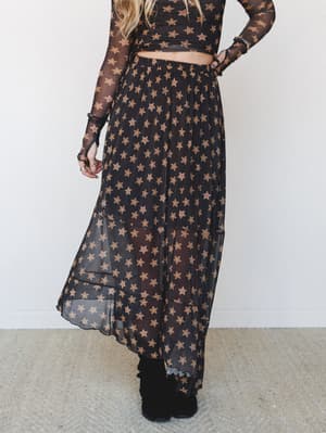 A sheer black maxi skirt covered in warm golden star print floats around the model's ankles, paired with a matching long-sleeve mesh crop top and black ankle boots — celestial and a little dramatic in the best way.