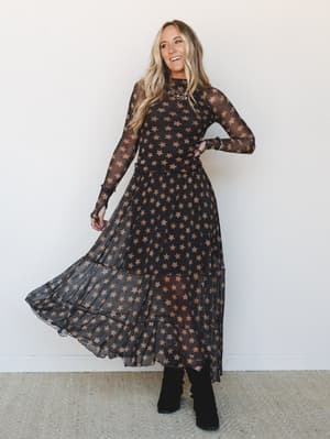 A smiling model twirls in a sheer black maxi dress covered in warm caramel stars, paired with black ankle boots and a statement necklace — the kind of outfit that makes a Tuesday feel like something worth dressing for.