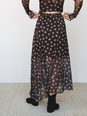 Shot from behind, a sheer black mesh maxi skirt with an allover warm-toned star print is styled with a matching long-sleeve crop top and black ankle boots — the kind of outfit that feels equally at home at a concert or a candlelit dinner.