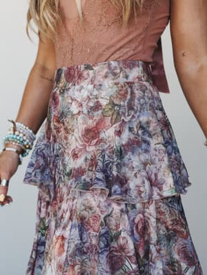 A close-up of a dreamy floral midi skirt in dusty rose and lavender, styled with a lace-back top and layered bracelets — the kind of outfit that looks like it belongs at a garden wedding or a very good afternoon.
