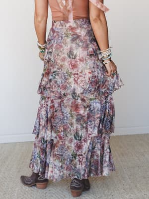 A tiered floral maxi skirt in soft dusty rose and muted lavender tones styled from the back with a blush tank, cowboy boots, and stacked bracelets — romantic and rooted at the same time.