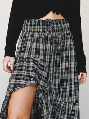A close-up of a flowy black plaid maxi skirt with a smocked waistband and ruffle-edged high-low hem, styled with a black long-sleeve crop top and stacked silver rings — relaxed but intentional, like a Saturday that turned into something.